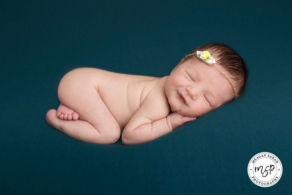 Baby girl newborn photography in Leeds. Photo on dark turquoise background smiling. Wearing headband