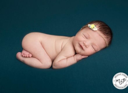 Baby girl newborn photography in Leeds. Photo on dark turquoise background smiling. Wearing headband