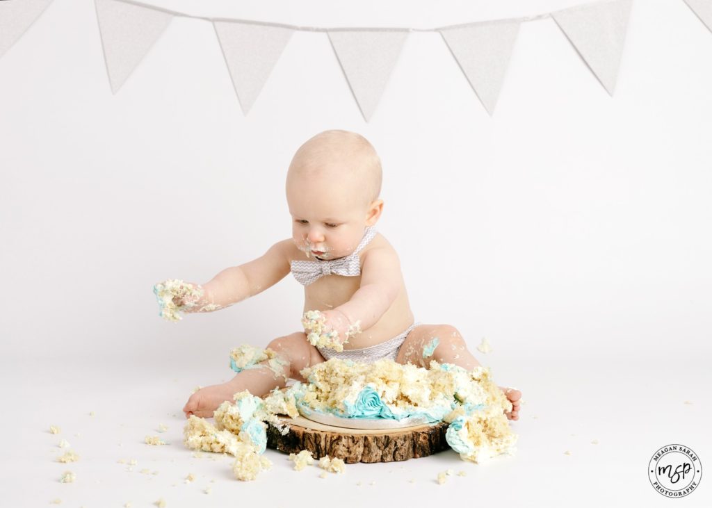 A photo of a baby in grey bow tie and nappy cover. Smashing a cake for his Cake Smash session in Leeds at Meagan Sarah Photography Studio. White background. Grey bunting. 