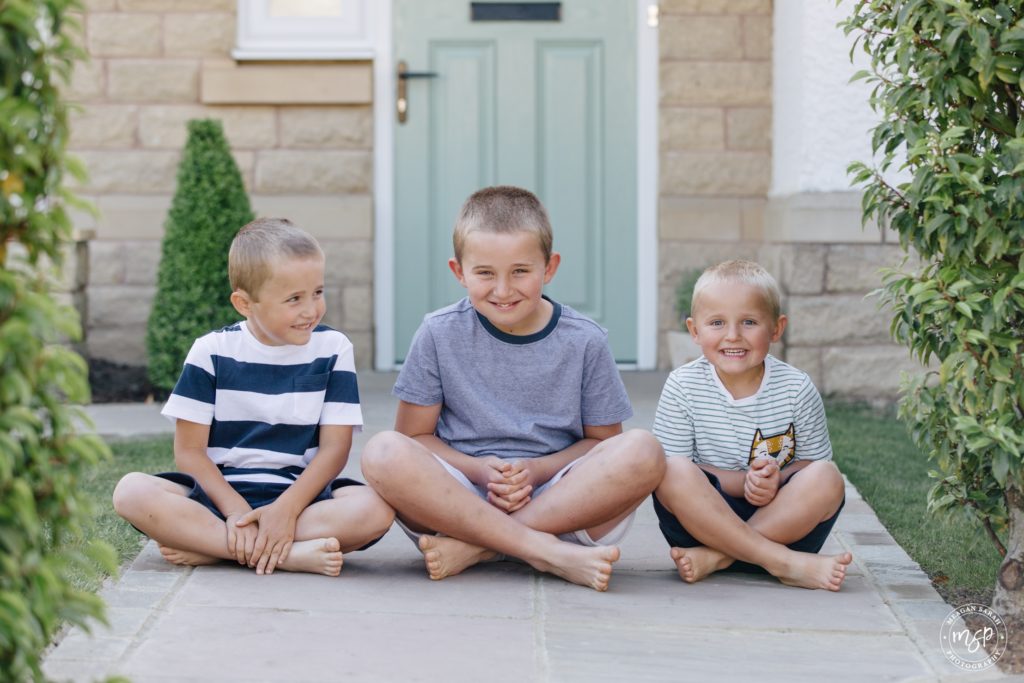 Photos of Doorstep Photography. Family of five outside their home. Picture of doorstep portrait in Horsforth, Leeds by professional photographer, Meagan Sarah Photography. Three boys sitting together, Brothers, During Coronavirus outbreak. Charity Photoshoot. Rerdrow Houses. Redrow homes. Yorkshire. Horsforth Vale.