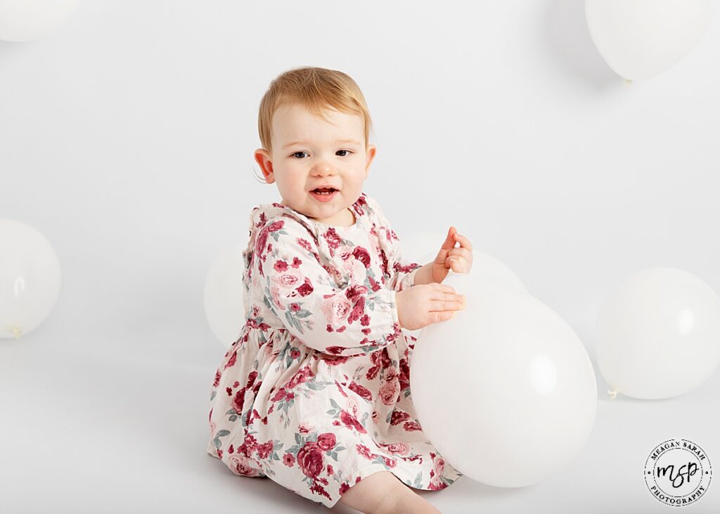 little girl on white background with white balloons in a pink floral dress facing camera smiling holding one loose balloon