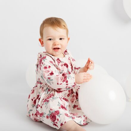 little girl on white background with white balloons in a pink floral dress facing camera smiling holding one loose balloon