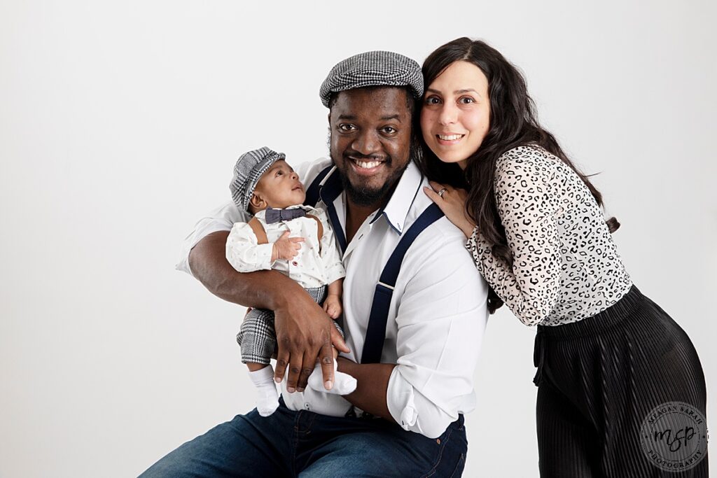 mum, dad and baby facing camera. dad and baby in flat caps and braces mum dressed in leopard print top and black trousers on a white background