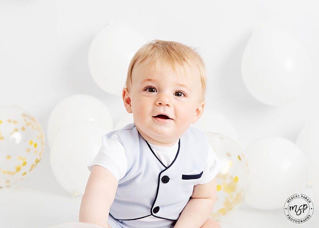 Smartly dressed little 1 year old boy sitting in front of white balloons, smiling at the camera.