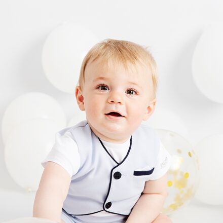 Smartly dressed little 1 year old boy sitting in front of white balloons, smiling at the camera.