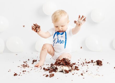 Little boy at his first birthday studio cake smash. Smashing his chocolate cake up and having fun. White set white balloons.