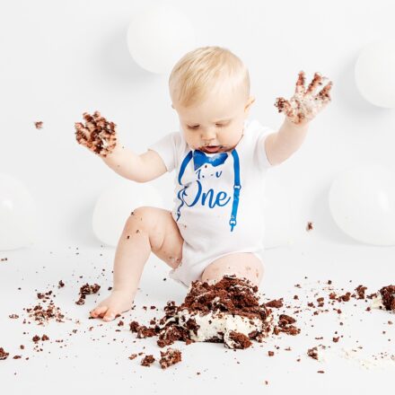 Little boy at his first birthday studio cake smash. Smashing his chocolate cake up and having fun. White set white balloons.