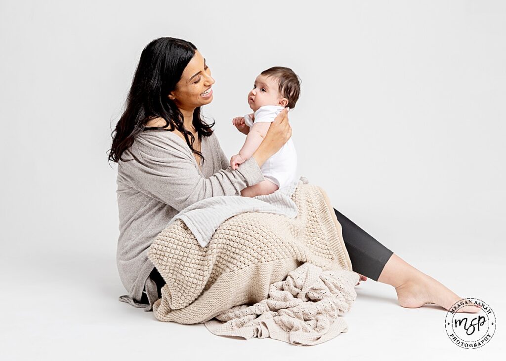 mum and 3 month old daughter sat on white background as mum hold baby up to her face with a smile