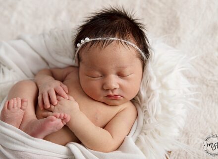 A beautiful, dark hair baby with headband on, laying on fur rug, asleep in a fruit bowl.
