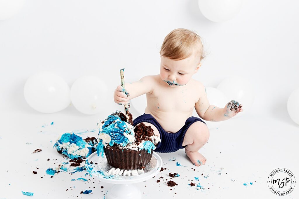 Little boy smashing a blue chocolate cake up with a wooden spoon at his cake smash photo session.