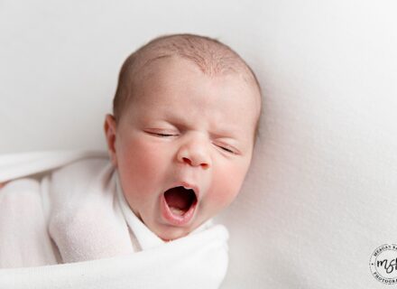 newborn baby boy on white background in white swaddle facing forwards yawning at the camera