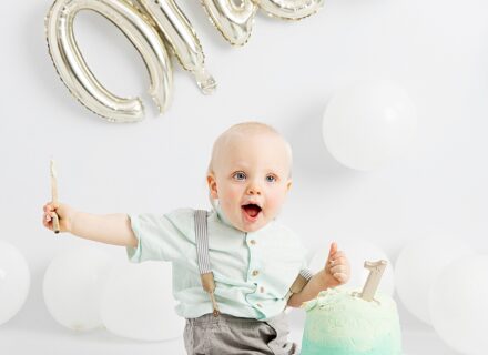 birthday boy one year old on white background with white balloons and inflatable silver one balloon with green birthday cake in the middle where he is sat