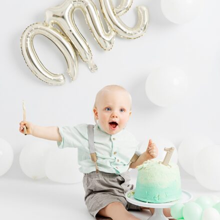 birthday boy one year old on white background with white balloons and inflatable silver one balloon with green birthday cake in the middle where he is sat
