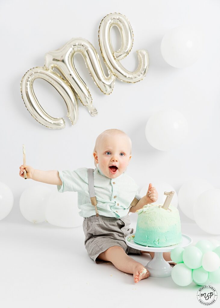 birthday boy one year old on white background with white balloons and inflatable silver one balloon with green birthday cake in the middle where he is sat