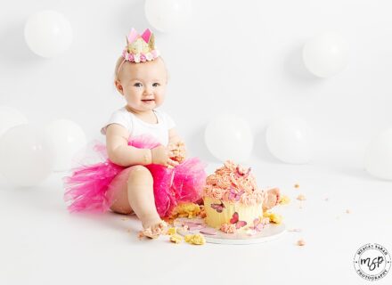 Cute little one year old baby girl at her studio cake smash session. Smiling at the camera with a pink tutu on and white balloons behind her.