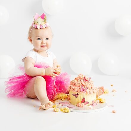 Cute little one year old baby girl at her studio cake smash session. Smiling at the camera with a pink tutu on and white balloons behind her.