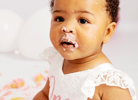 little girl with pigtails facing the camera with pink icing and cake on her face around nose and mouth in a birthday dress saying one in pink