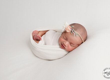 newborn baby girl on white background swaddled in white fabric with a delicate rose head band on with a slight smile on her face