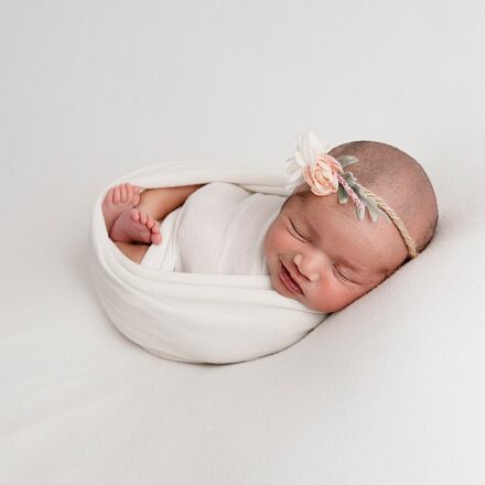 newborn baby girl on white background swaddled in white fabric with a delicate rose head band on with a slight smile on her face