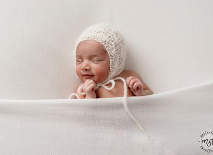 baby girl on what background wearing white knitted bonnet tied under chin with a white blanket over chest and hands raised above it