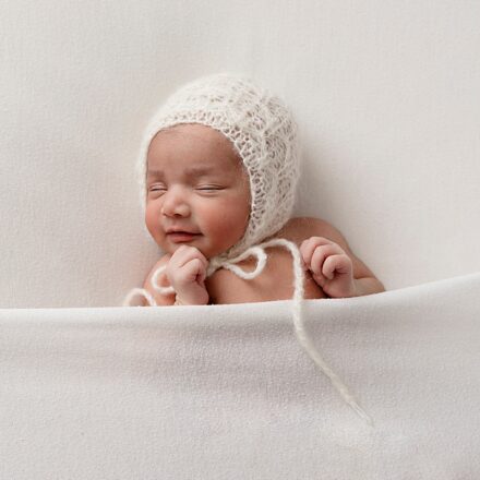 baby girl on what background wearing white knitted bonnet tied under chin with a white blanket over chest and hands raised above it