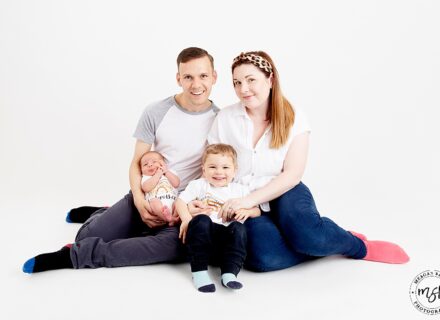 family photo shoot on a white background including mum dad newborn baby and toddler brother. all wearing white tops and blue trousers smiling at the camera