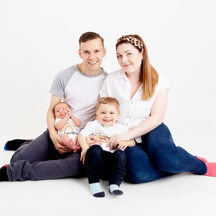 family photo shoot on a white background including mum dad newborn baby and toddler brother. all wearing white tops and blue trousers smiling at the camera