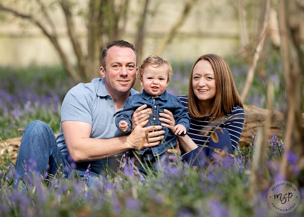Cute little boy smiling with his mum and dad. sitting in the woods among bluebells for their photoshoot. Wearing blue denim. Wakefield.