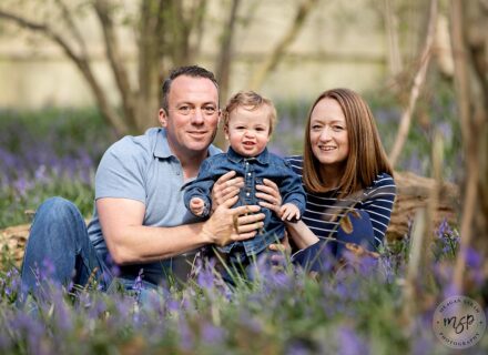 Cute little boy smiling with his mum and dad. sitting in the woods among bluebells for their photoshoot. Wearing blue denim. Wakefield.