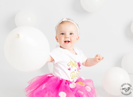 little girl, on white set with white balloons in a pink tutu and white headband facing the camera holding a balloon and smiling
