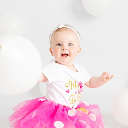 little girl, on white set with white balloons in a pink tutu and white headband facing the camera holding a balloon and smiling