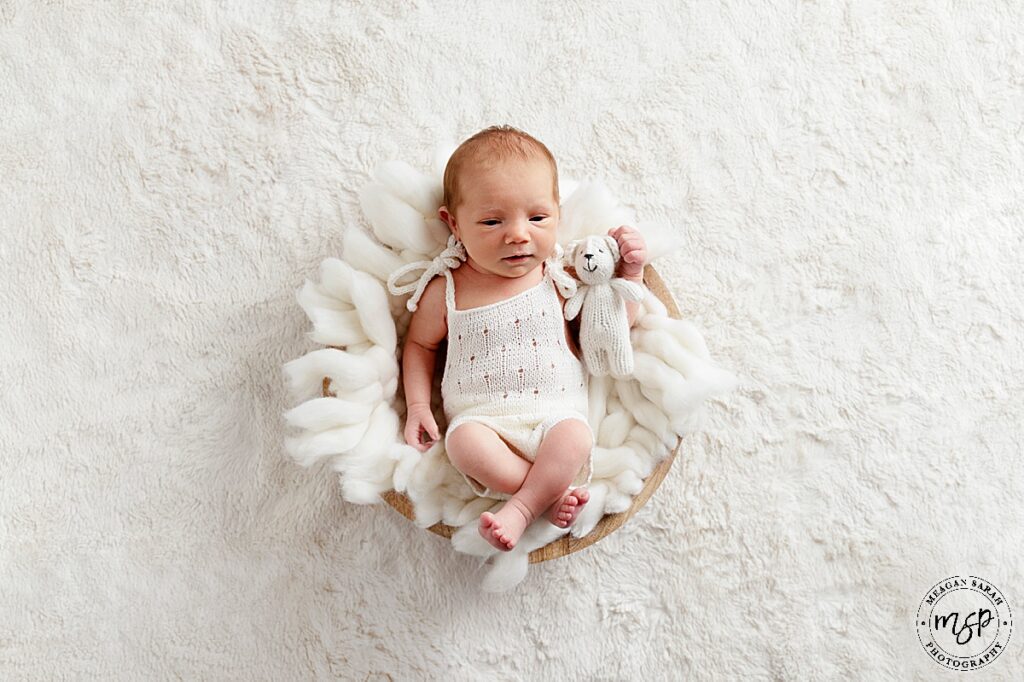 baby girl in a basket with white wool and w cream rug underneath in a cream romper looking at the camera