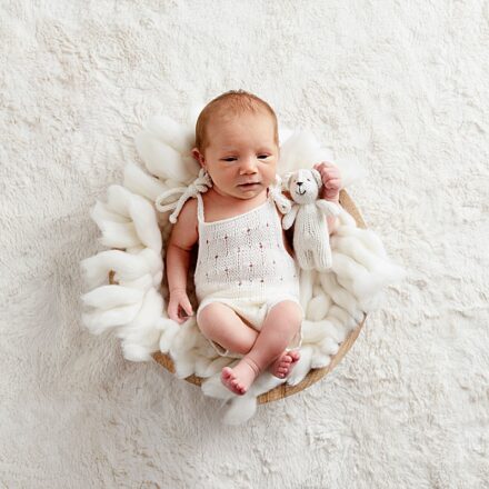 baby girl in a basket with white wool and w cream rug underneath in a cream romper looking at the camera