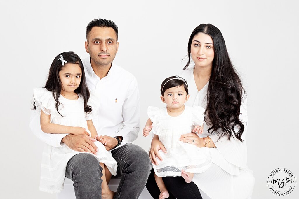 family of four on white backdrop all facing the camera smiling two children on either mum and dads knee dressed in white