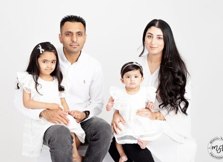 family of four on white backdrop all facing the camera smiling two children on either mum and dads knee dressed in white