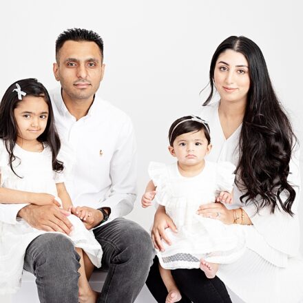 family of four on white backdrop all facing the camera smiling two children on either mum and dads knee dressed in white