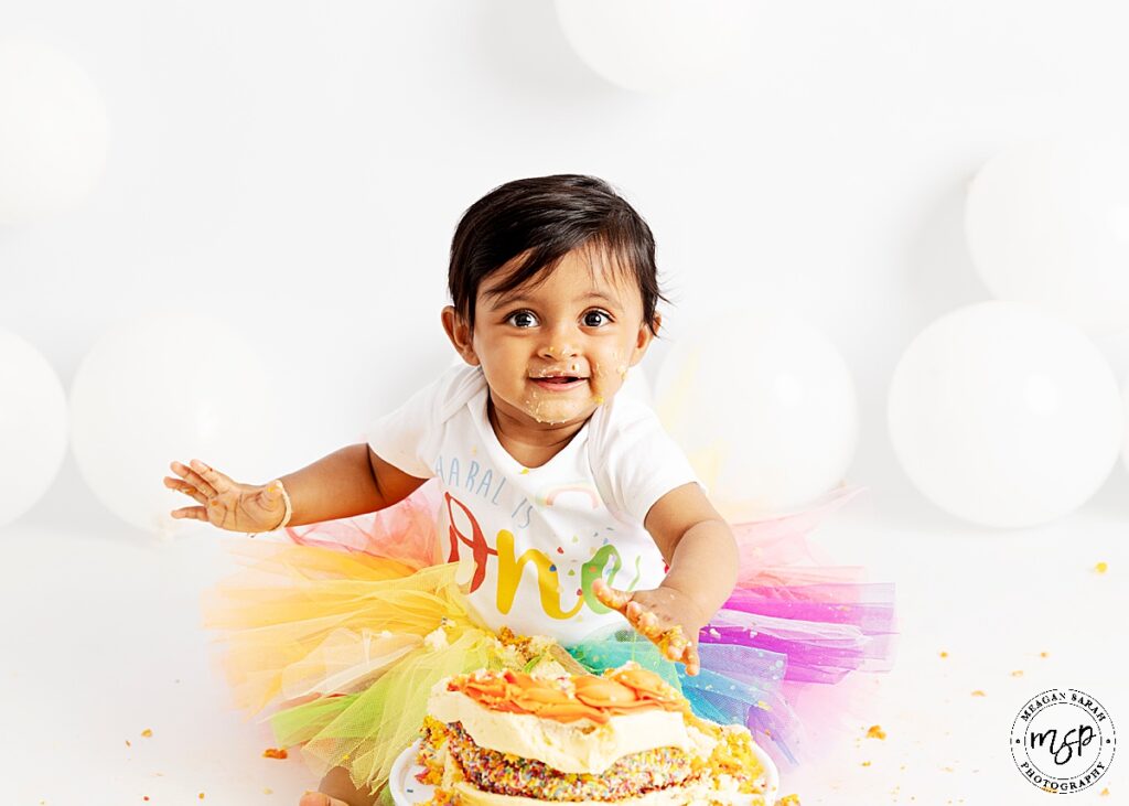 little girl in rainbow tutu with white baby grow on saying ONE facing the camera smiling with cake in her hand