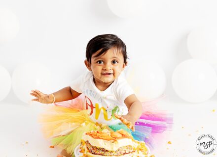 little girl in rainbow tutu with white baby grow on saying ONE facing the camera smiling with cake in her hand