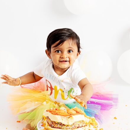 little girl in rainbow tutu with white baby grow on saying ONE facing the camera smiling with cake in her hand