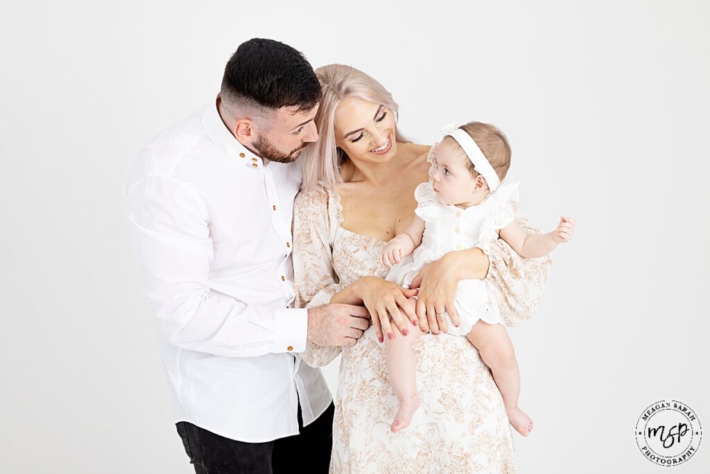 mum and dad and 3 month little girl all dressed in white neutral colours. mum holding little girl in arms looking down at her smiling