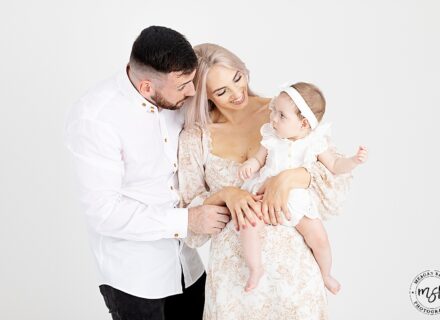 mum and dad and 3 month little girl all dressed in white neutral colours. mum holding little girl in arms looking down at her smiling