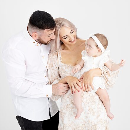mum and dad and 3 month little girl all dressed in white neutral colours. mum holding little girl in arms looking down at her smiling