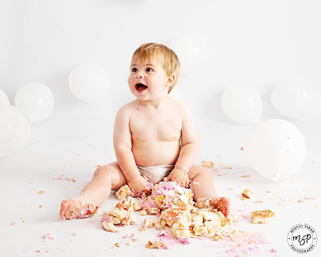 little boy sat against white backdrop with white balloon and a smash pink cake between his legs smiling off camera left