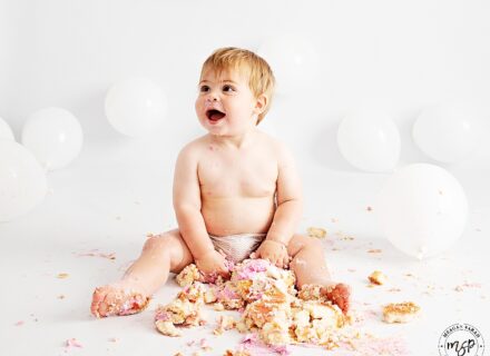 little boy sat against white backdrop with white balloon and a smash pink cake between his legs smiling off camera left