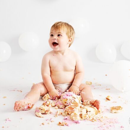 little boy sat against white backdrop with white balloon and a smash pink cake between his legs smiling off camera left