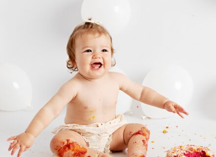 one year old girl sat on white background with white balloons in just cream shorts sat in front of a smashed rainbow cake. she is smiling