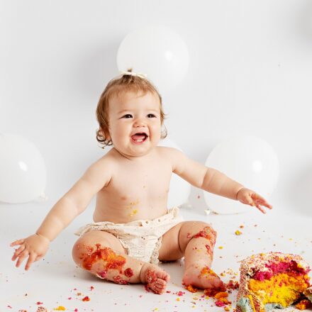 one year old girl sat on white background with white balloons in just cream shorts sat in front of a smashed rainbow cake. she is smiling