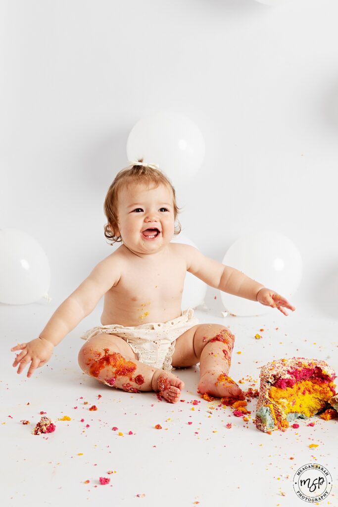 one year old girl sat on white background with white balloons in just cream shorts sat in front of a smashed rainbow cake. she is smiling