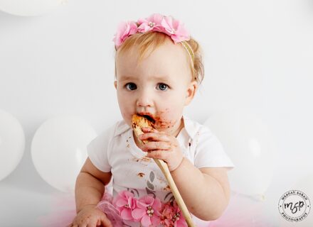 little girl on white backdrop dressed in white and pink with pink headband, holden spoon and licking chocolate off it