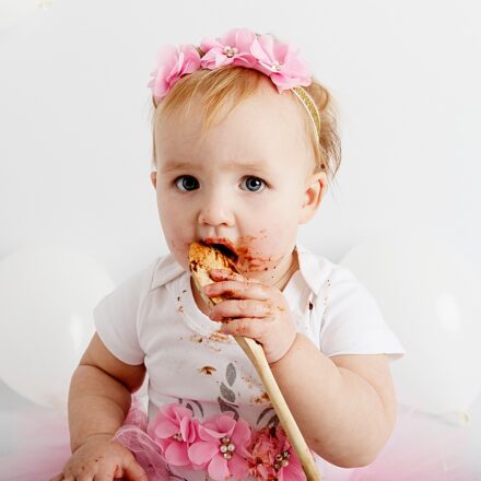 little girl on white backdrop dressed in white and pink with pink headband, holden spoon and licking chocolate off it